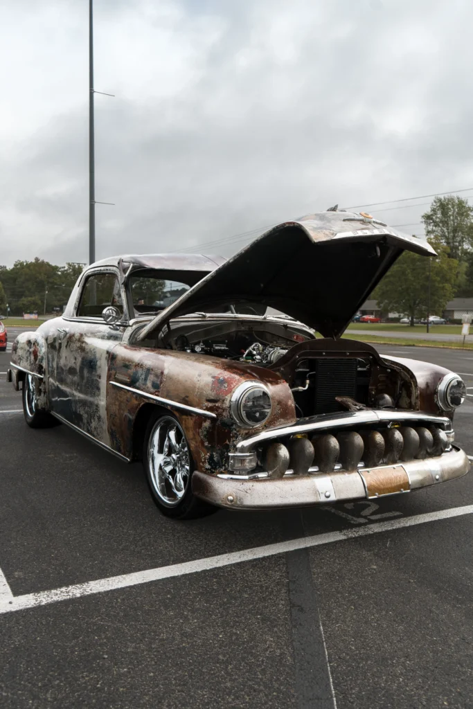 Rusted and patina-covered Plymouth Cambridge with hood open in a parking lot under cloudy sky