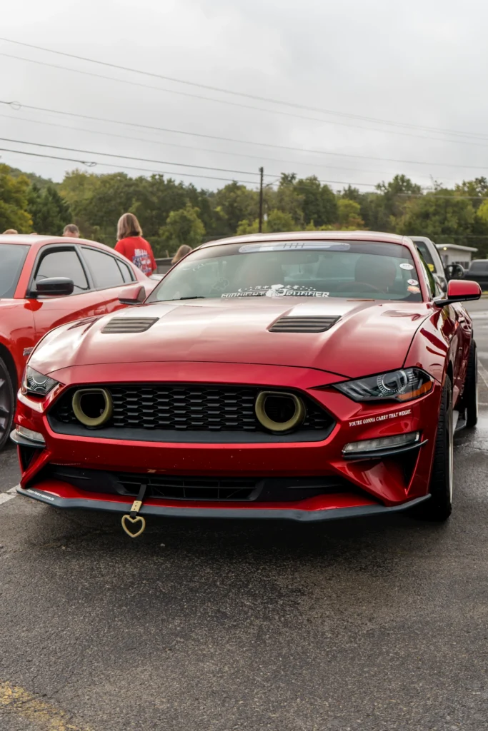 Red Ford Mustang with aftermarket modifications parked outdoors with another red vehicle and people in the background