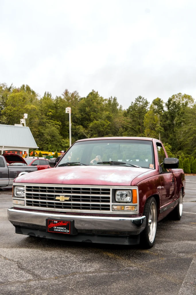 A burgundy Chevrolet C/K pickup with faded hood paint is parked at an outdoor car event.