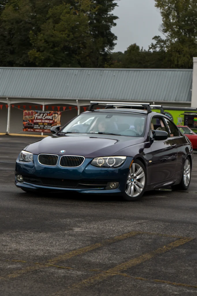 Dark blue BMW 3 Series coupe with roof rack parked in lot, building and trees in background