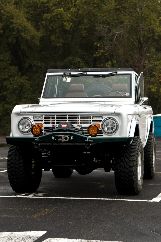 White Ford Bronco parked in front of green foliage, showing front view with aftermarket lights and winch