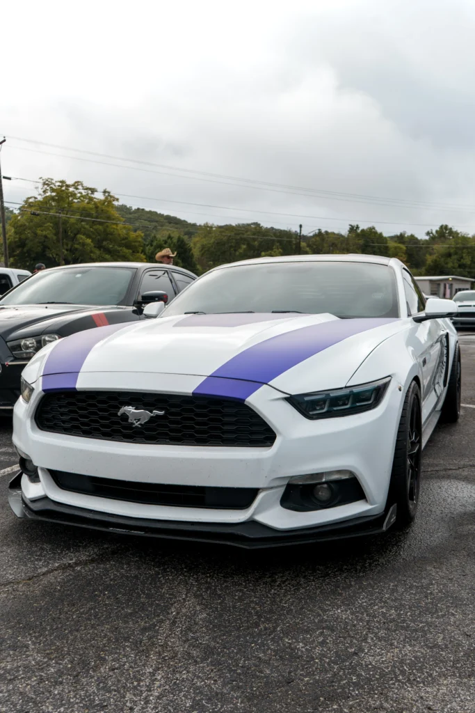 White Ford Mustang with blue racing stripes parked outdoors with another black car nearby and trees in the background