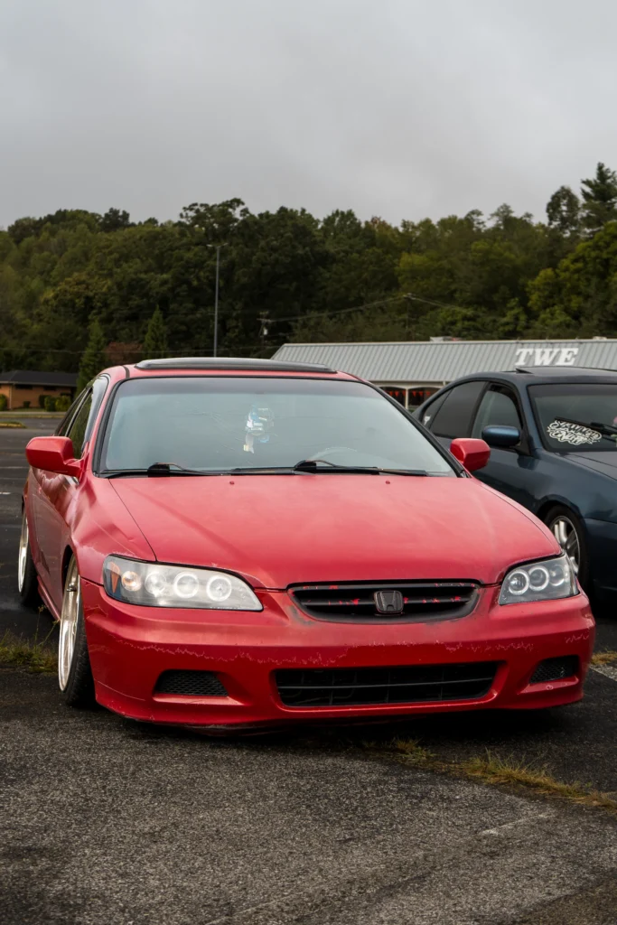 A red Honda Accord with worn paint and a cracked windshield is parked beside another car in an outdoor lot.