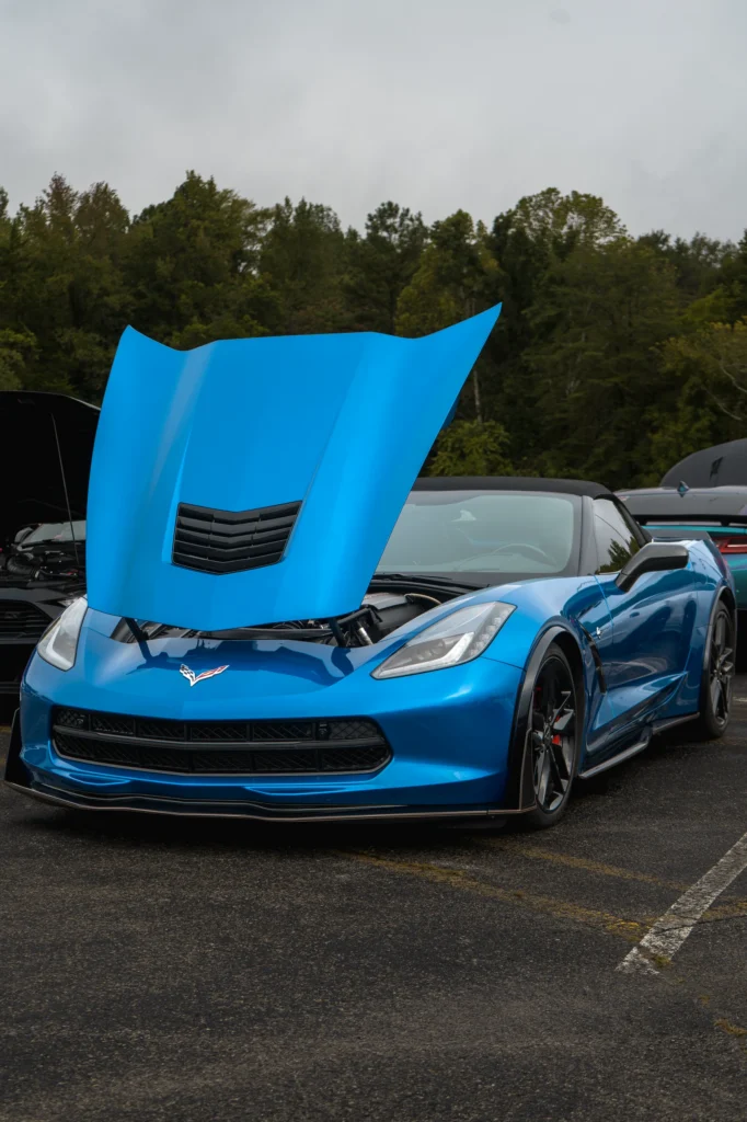 Blue Chevrolet Corvette with hood open at an outdoor car event, surrounded by other vehicles and trees in background.