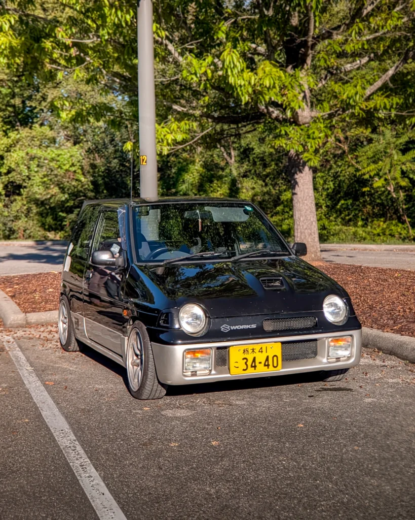 Black Suzuki Alto Works parked in a lot with trees in the background
