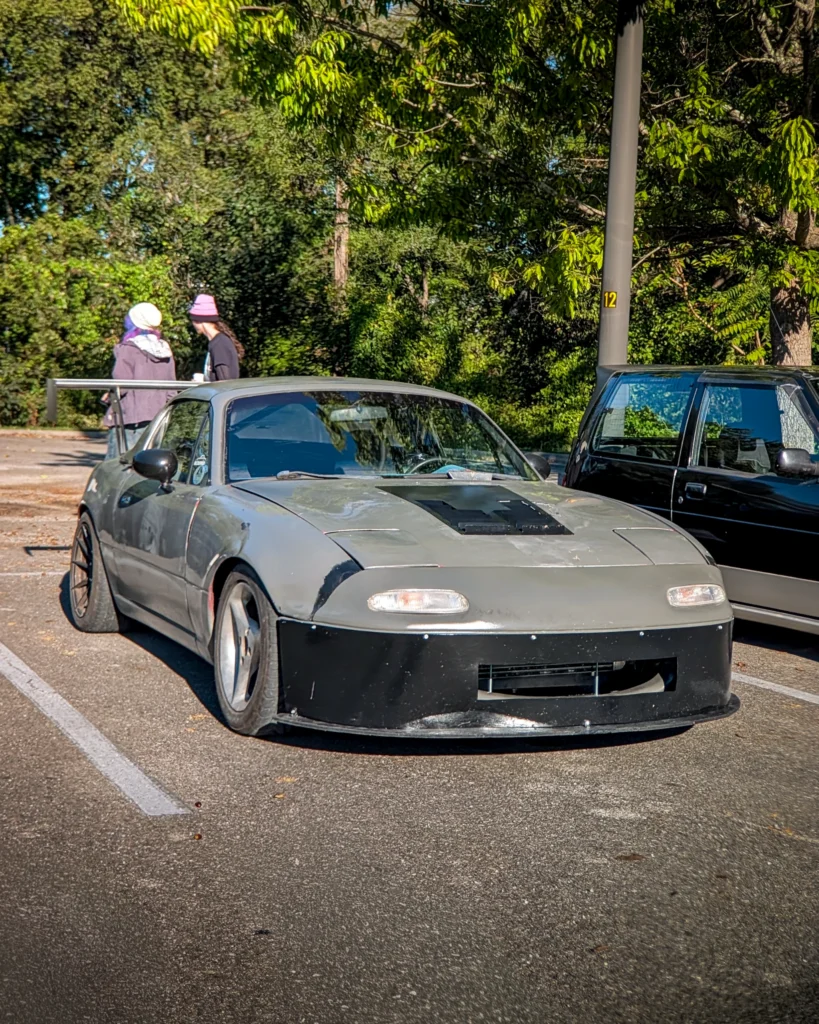 Gray Mazda MX-5 Miata with large rear wing parked in a lot with two people walking in the background