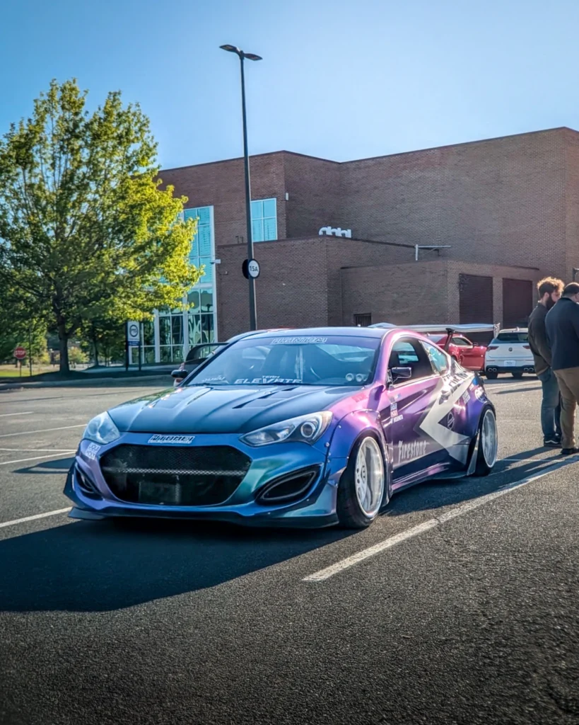 A blue and purple Hyundai Genesis Coupe with widened fenders is parked beside attendees in an outdoor lot.