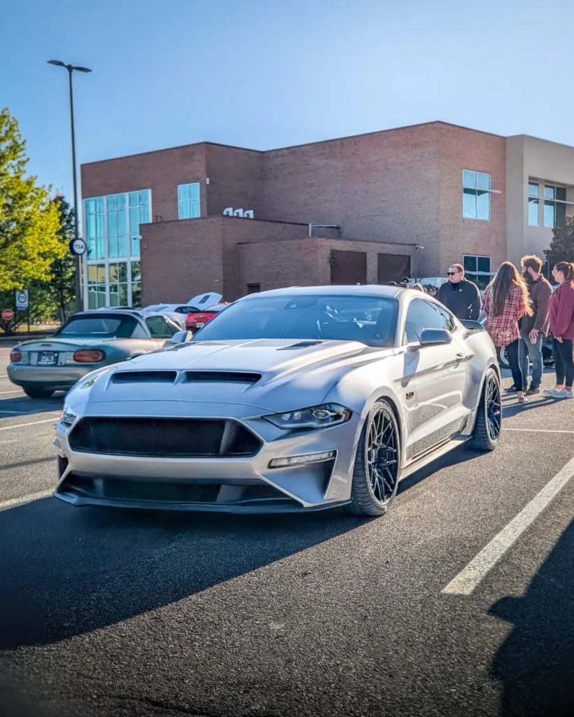 Silver Ford Mustang GT parked in a lot with people standing nearby and other cars in background by brick building