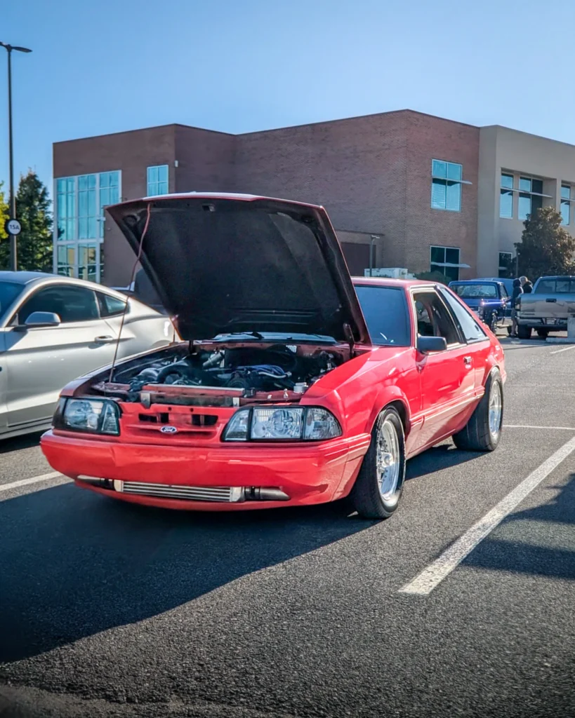 A red Ford Mustang sits with its hood open in a parking lot beside other vehicles at a car show.