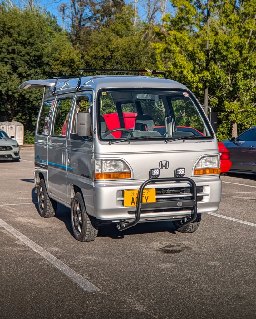A silver Honda Acty van with a roof rack and front brush guard is parked among other cars in a tree-lined lot.