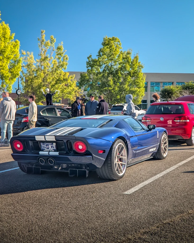 Blue Ford GT sports car with white racing stripes parked in a lot with several people nearby and other cars around.