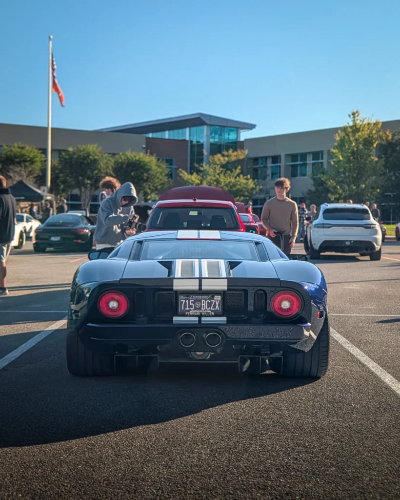 Black Ford GT with white racing stripes parked in a lot with other cars and people around, under clear sky.