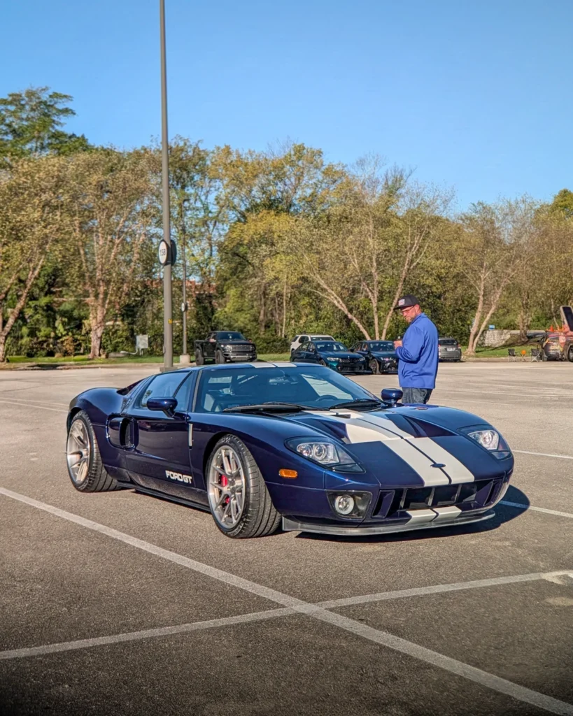 Dark blue Ford GT with white racing stripes parked in a lot with trees and other vehicles in the background