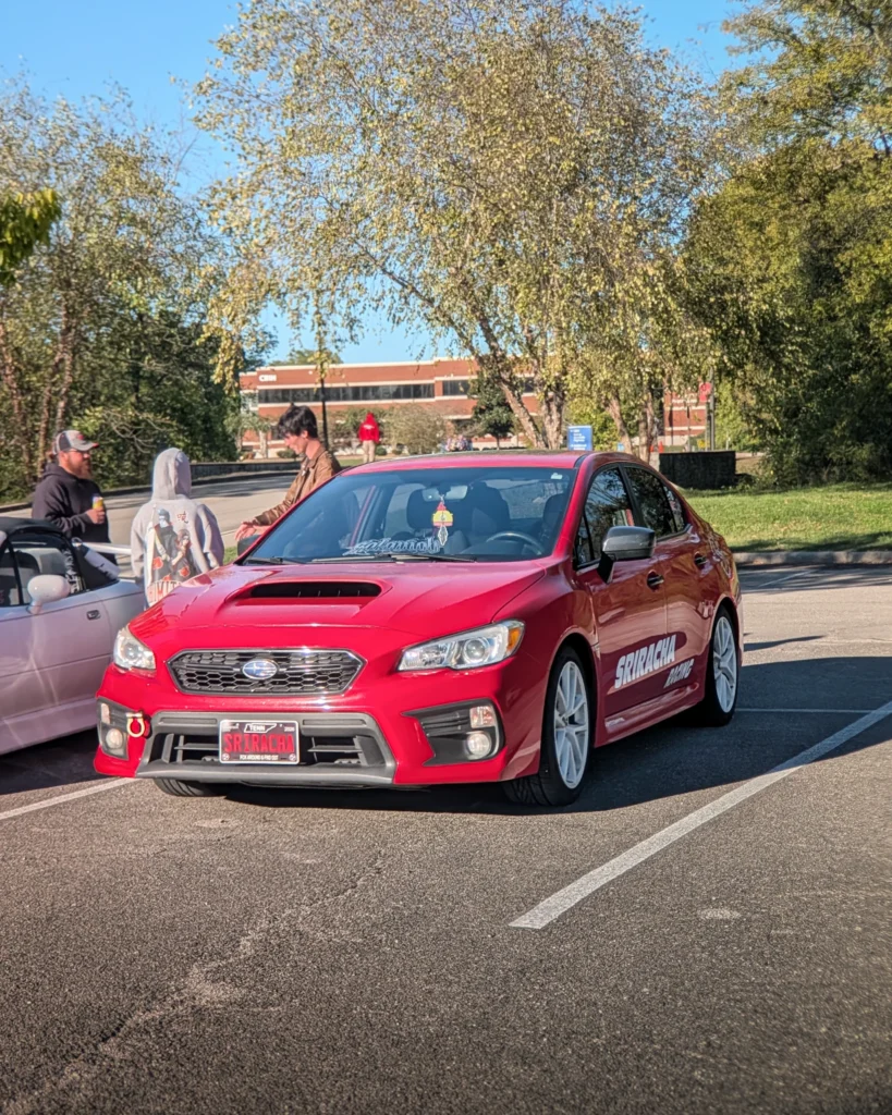 Red Subaru WRX sedan parked at a car event with several people nearby and a white convertible in background