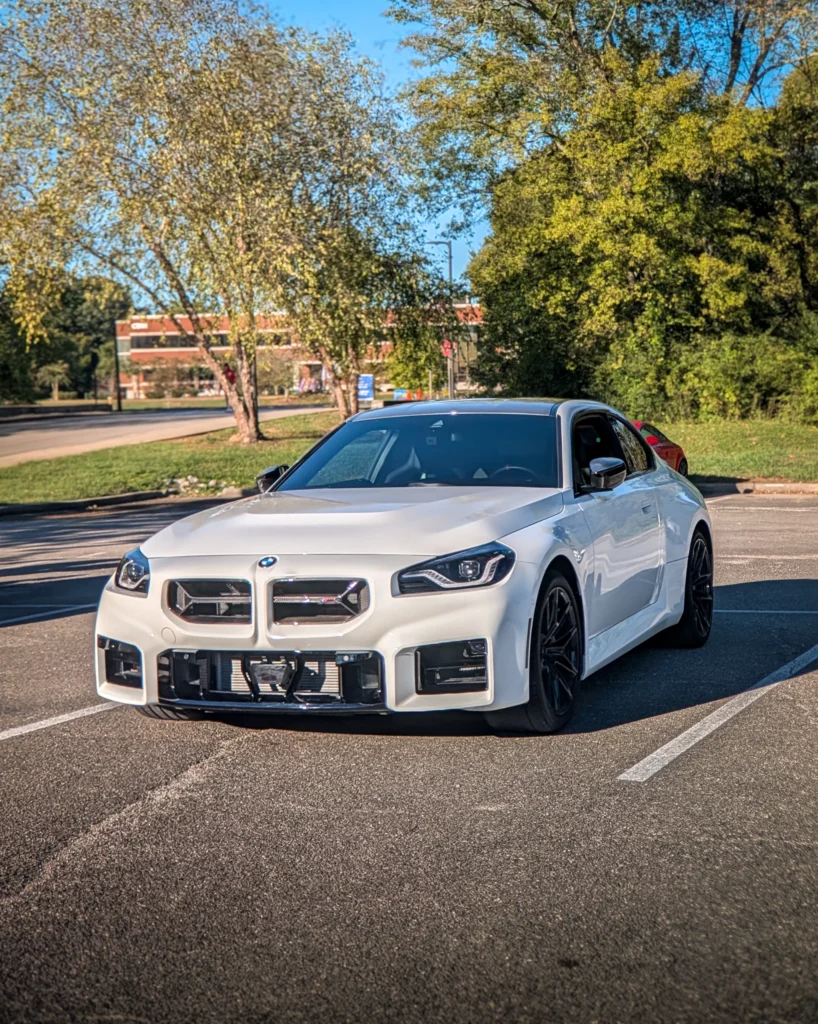 A white BMW M2 coupe is parked in a marked asphalt lot beside trees, viewed from the front three-quarter angle.