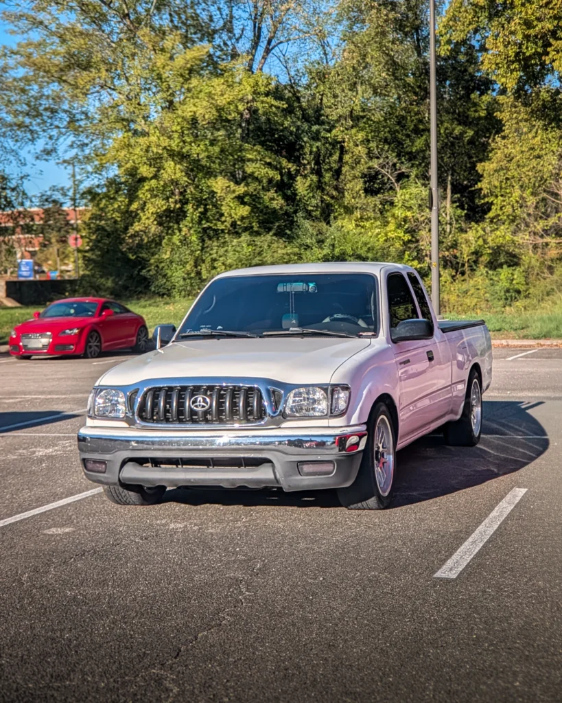 White Toyota Tacoma pickup truck parked in a lot with a red Audi TT in the background and trees behind