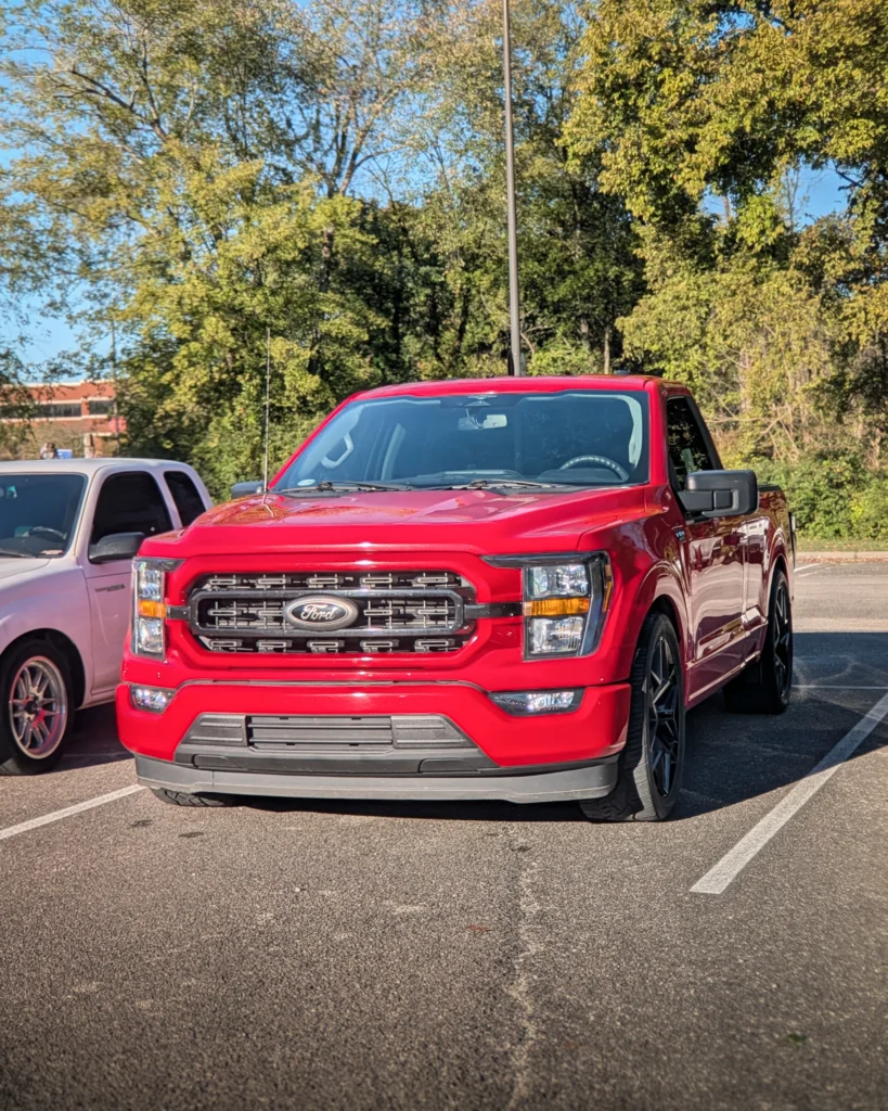 A red Ford F-150 is parked beside another pickup in a tree-lined parking lot.