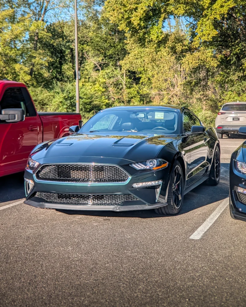 Dark green Ford Mustang parked beside a red pickup truck and behind it a white Mazda CX-5 with trees in background