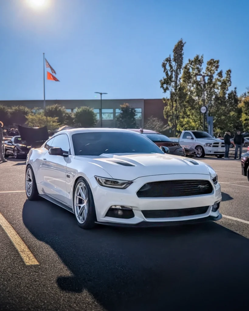 A white Ford Mustang parked at an outdoor car show, with other vehicles and attendees behind it.