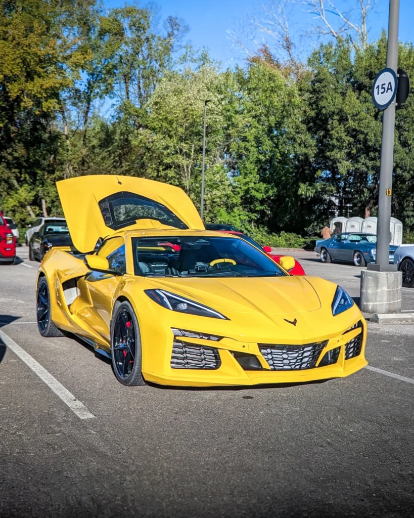 Yellow Chevrolet Corvette with rear hatch open parked outdoors at a car show with other vehicles and trees in the background