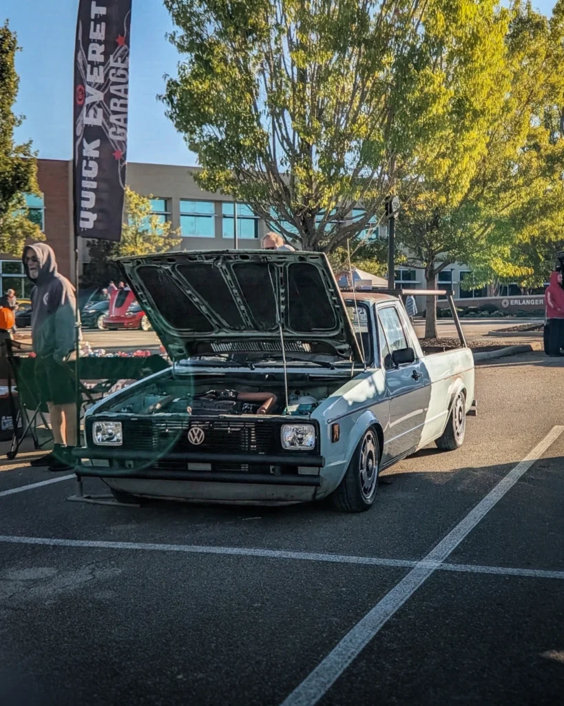 A light blue Volkswagen Rabbit pickup sits with its hood open in a parking lot at an outdoor car show.