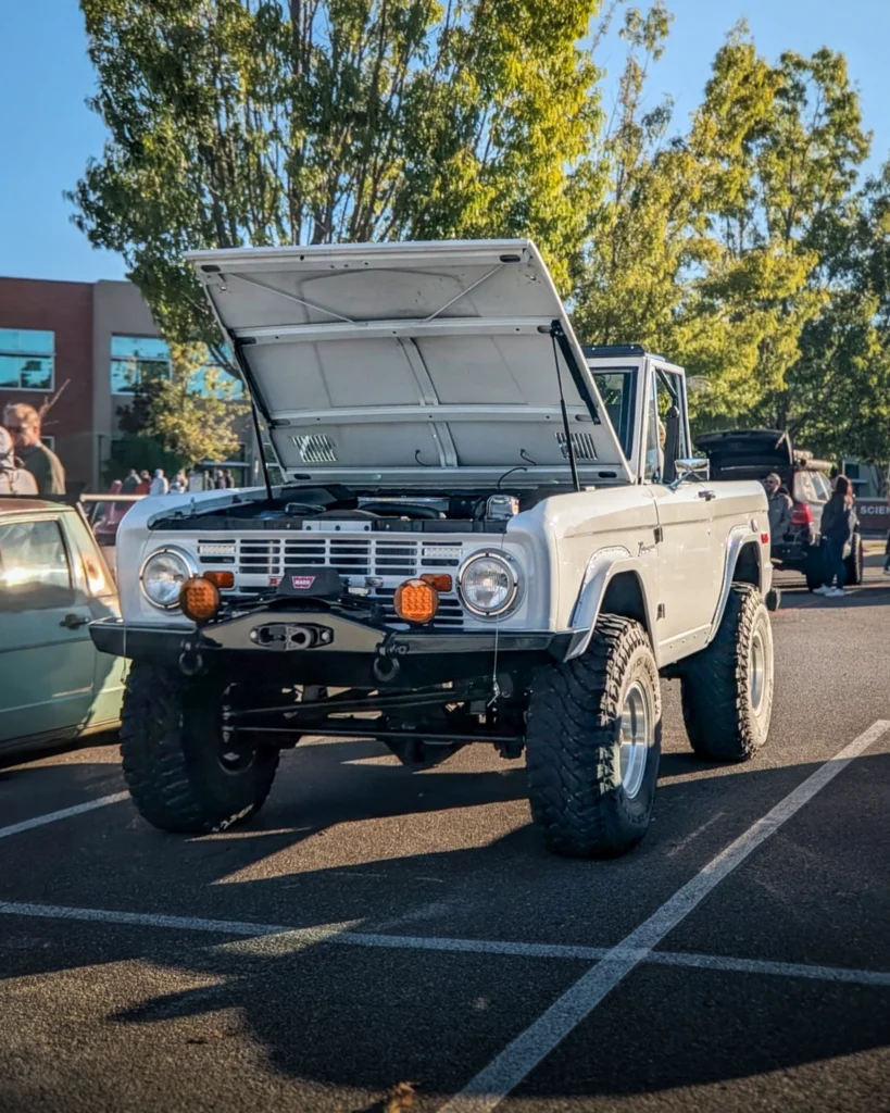 White Ford Bronco with its hood open at a car event parking lot with people and trees in the background.