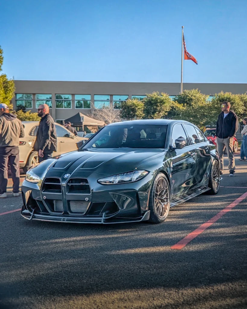 A dark green BMW M3 sits in a parking lot among attendees and other cars at an outdoor car event.