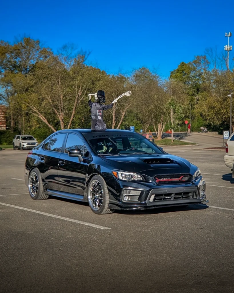Black Subaru WRX sedan with a Darth Vader skeleton figure on the roof in a parking lot with trees in background
