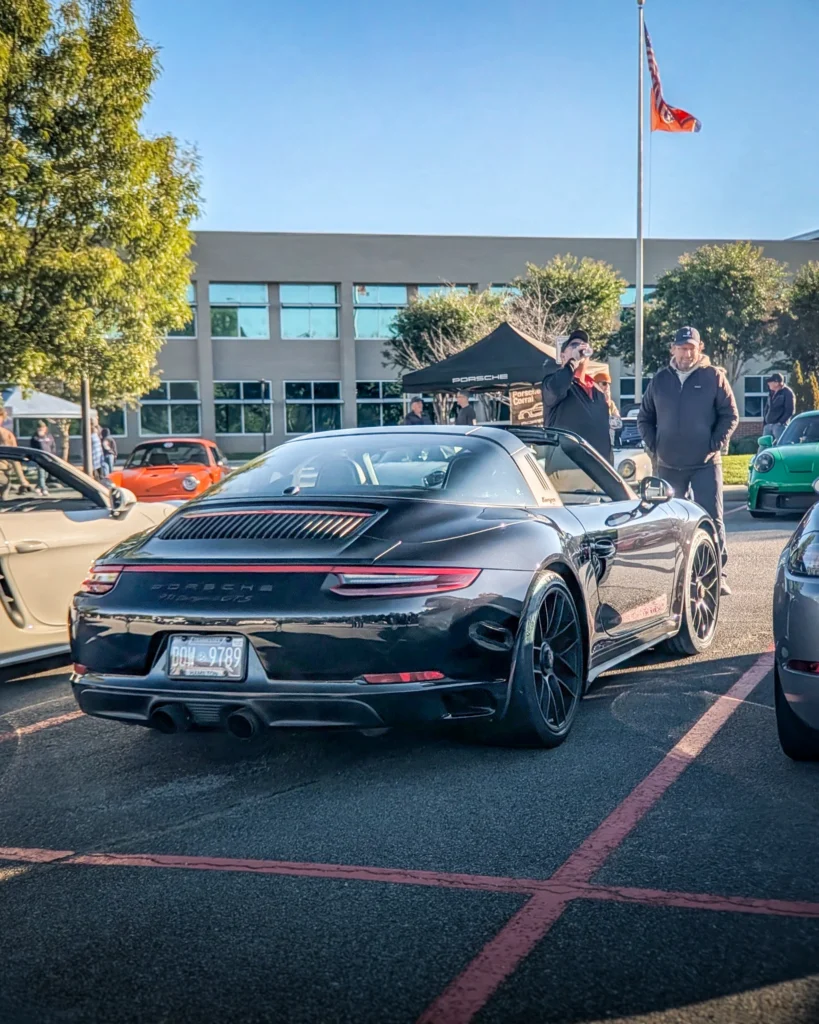 Black Porsche 911 Targa 4 GTS rear in a parking lot at a car event with people and tents in the background.