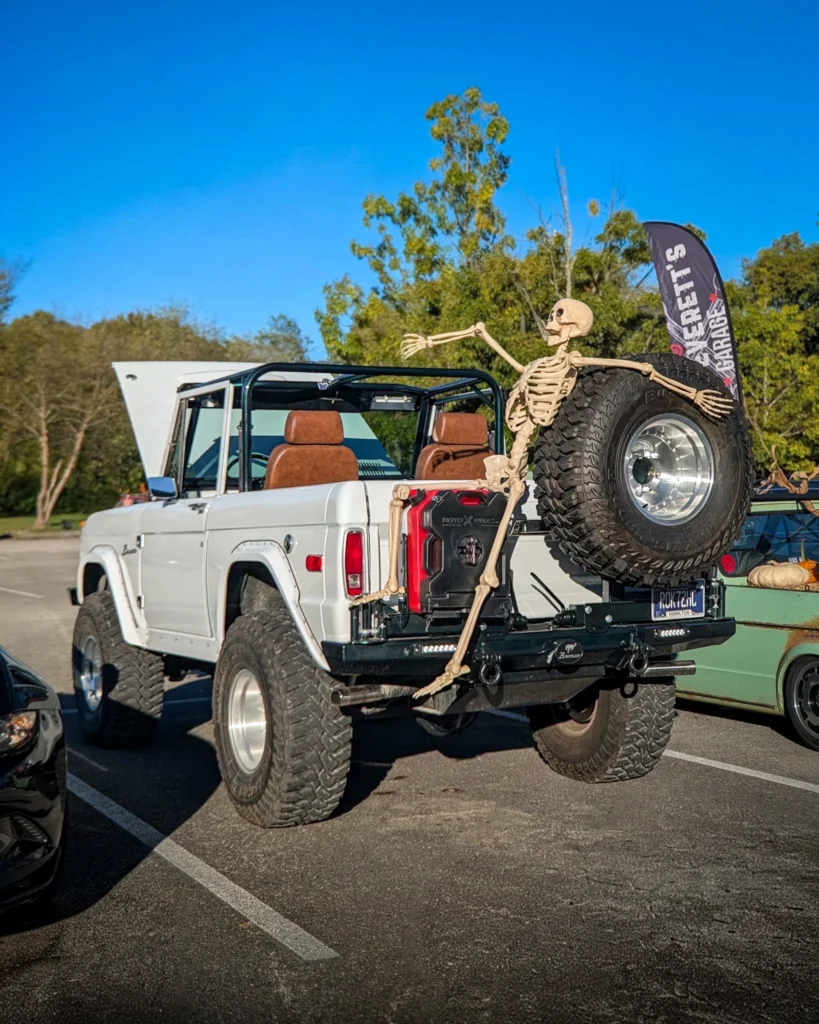 White Ford Bronco with brown seats and skeleton seated on spare tire in parking lot with trees and banner in background
