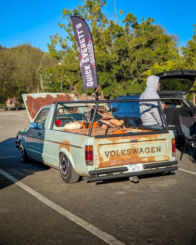 A light green Volkswagen Rabbit pickup with rusted panels and pumpkins in its bed sits at an outdoor car show.
