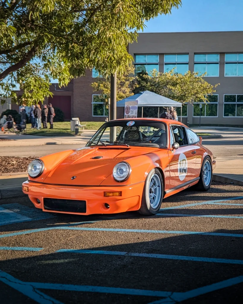 Orange Porsche 911 with racing number 12 parked outdoors near trees and a building with people in the background