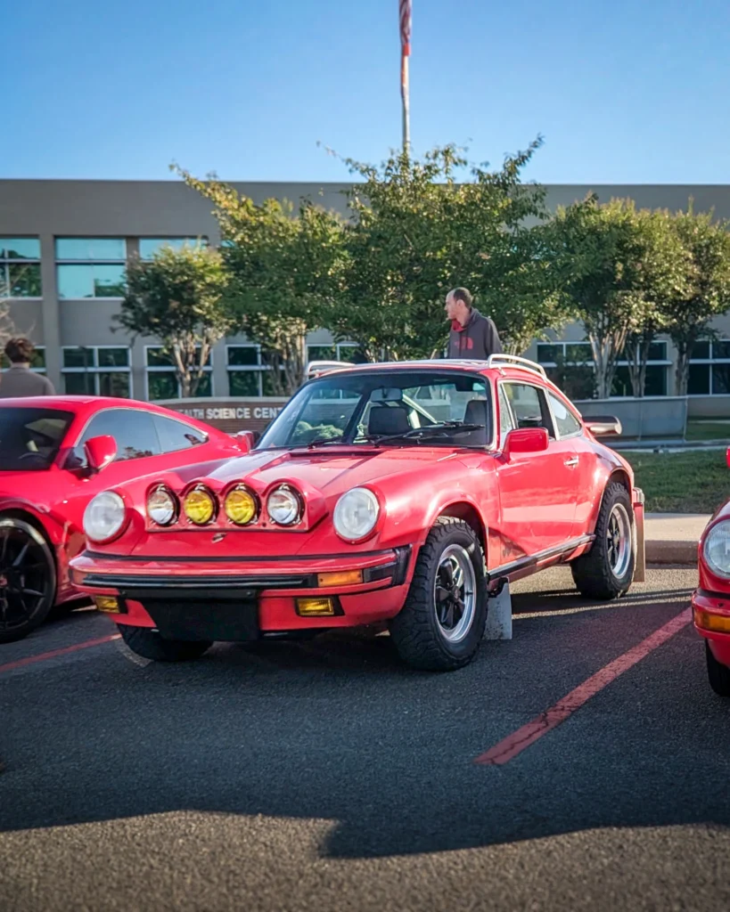 Red Porsche 911 with rally-style auxiliary lights and off-road tires parked next to a red coupe in a parking lot