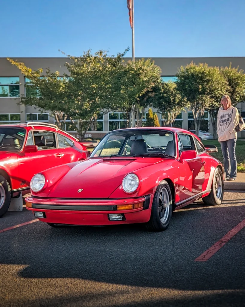 Red Porsche 911 coupe and red coupe parked outdoors near a person and trees in a parking lot