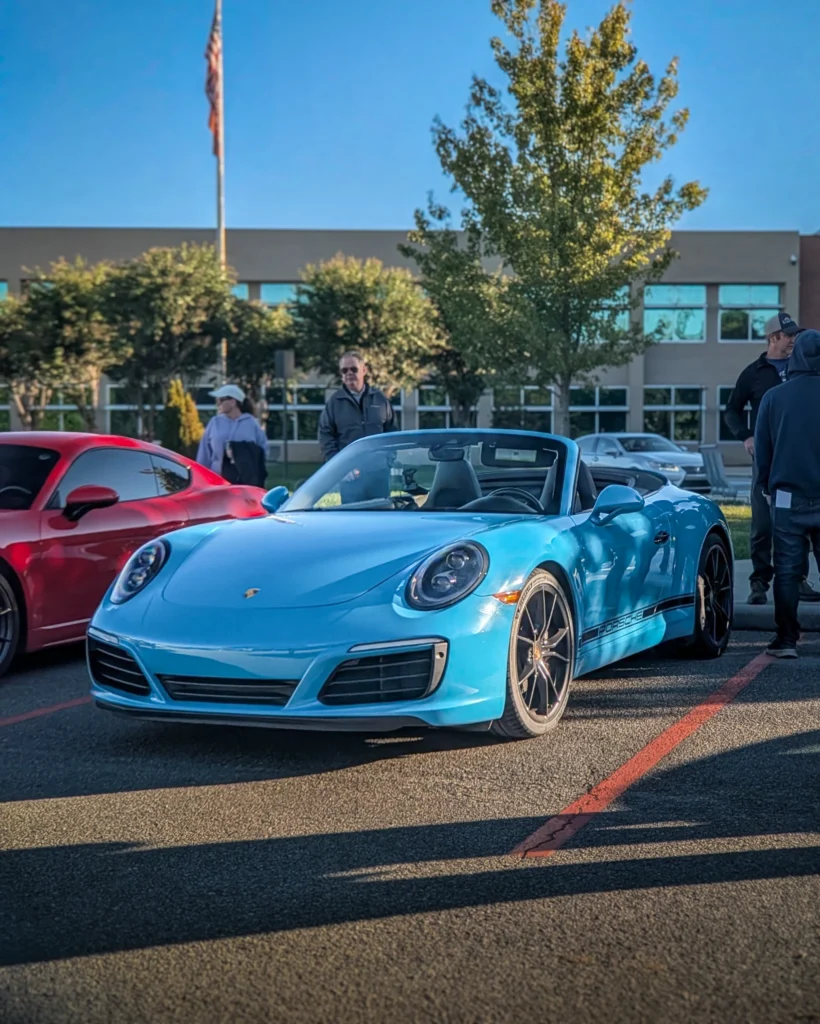 Blue Porsche 911 convertible and red coupe parked with people and trees in background at a car event