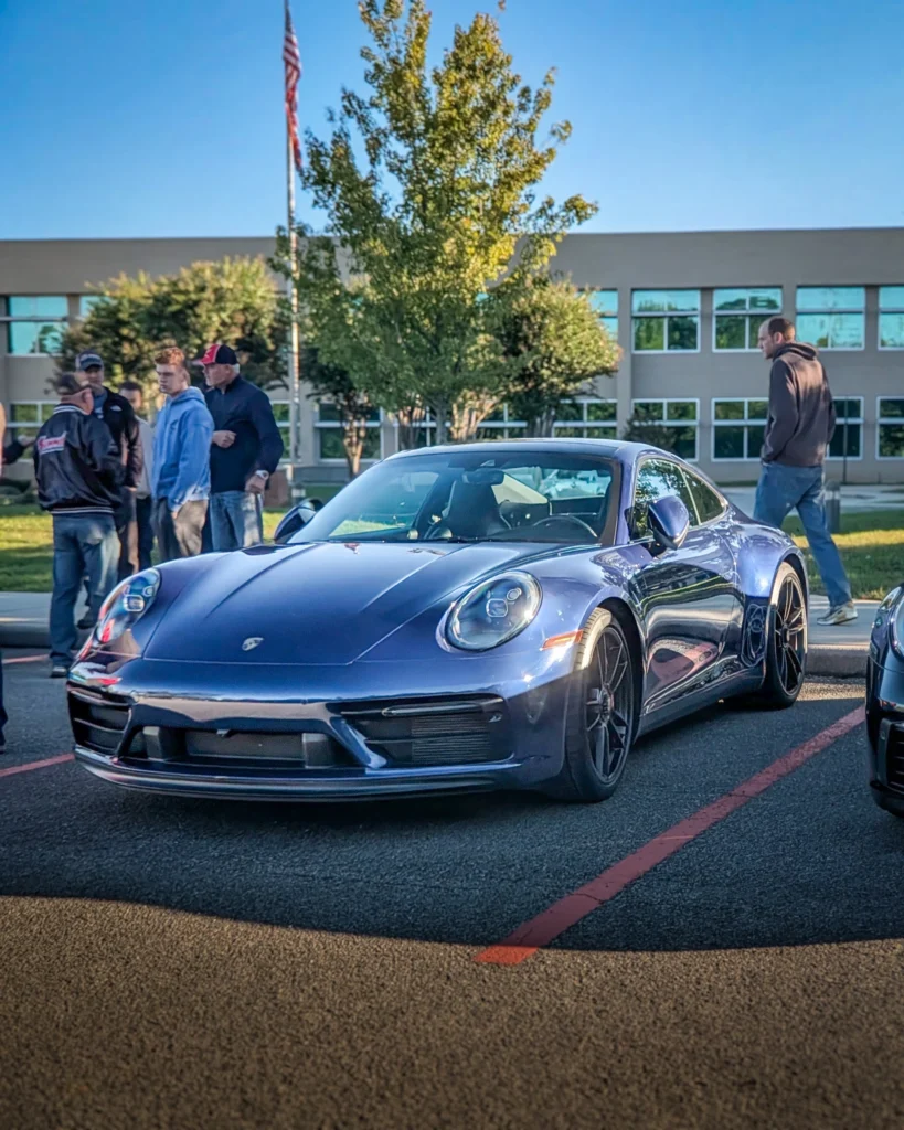 Blue Porsche 911 parked in a lot with people standing nearby and a building in the background under clear skies.