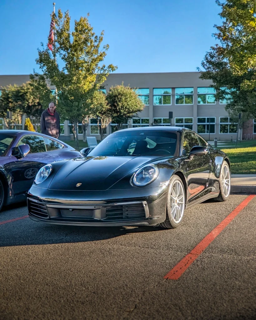 Black Porsche 911 parked outdoors next to a purple Porsche, with a building and trees in the background and a man walking