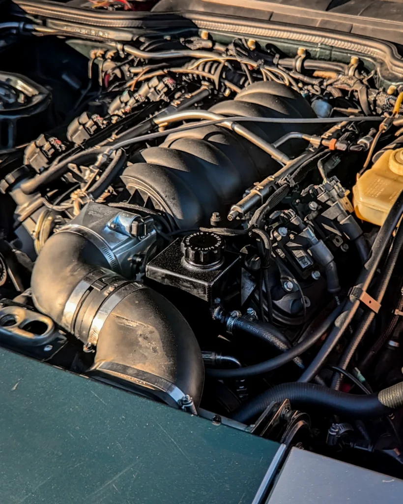 Close-up view of a Porsche 944 engine bay showing intake manifold, wiring, hoses, and fluid reservoir under sunlight.