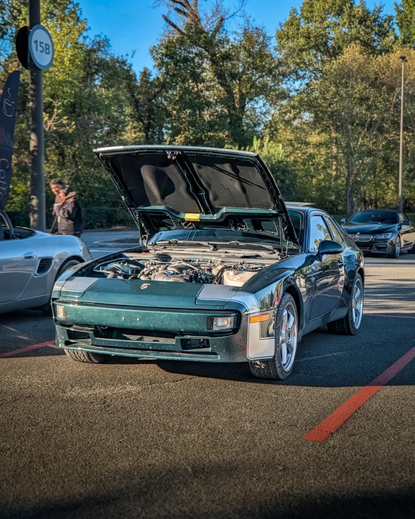 Green Porsche 944 with open hood parked on asphalt with trees and other cars in background