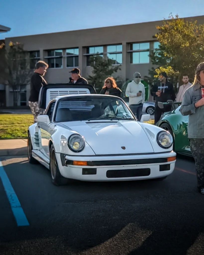 White Porsche 911 with rear engine lid open parked near building with several people in the background