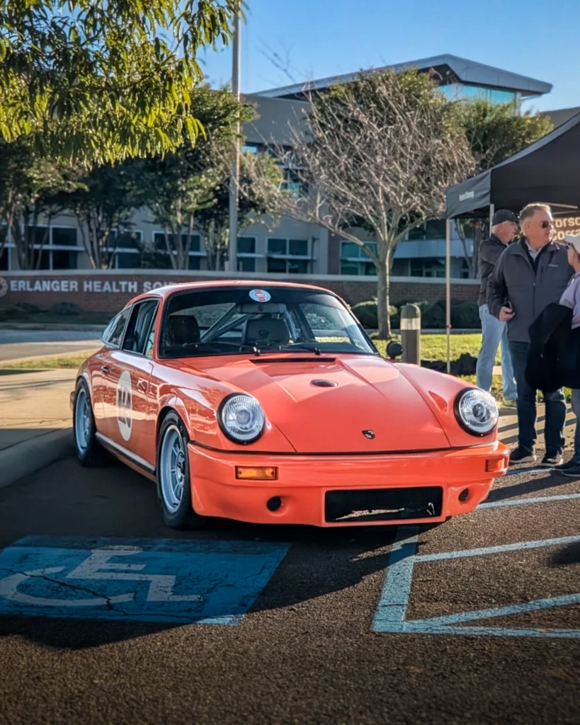 Orange Porsche 911 parked at an outdoor event with people and a building in the background