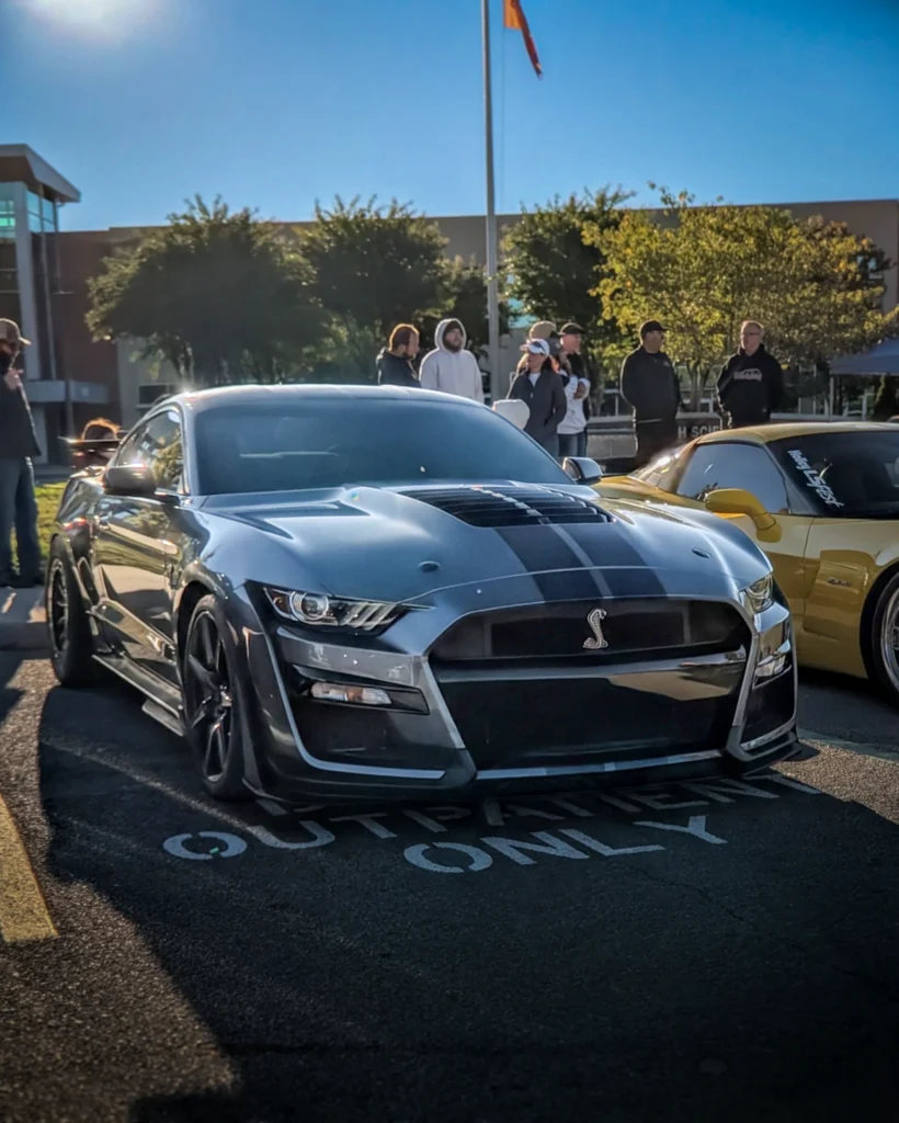 A silver Ford Mustang with black hood stripes is parked at an outdoor car show, with attendees behind it.