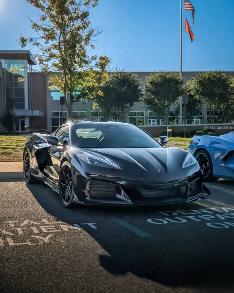 Black Chevrolet Corvette parked in front of a building with trees and flags in the background under a blue sky