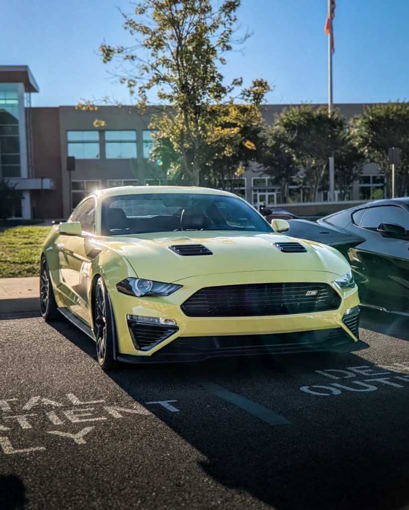 Yellow Ford Mustang parked outside under sunlight with building and trees in background