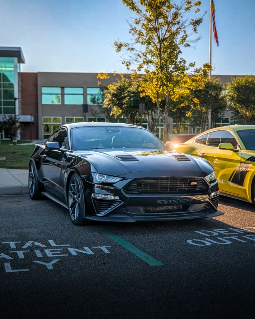 Black Ford Mustang and yellow Ford Mustang parked side by side in front of a modern building and trees at sunset