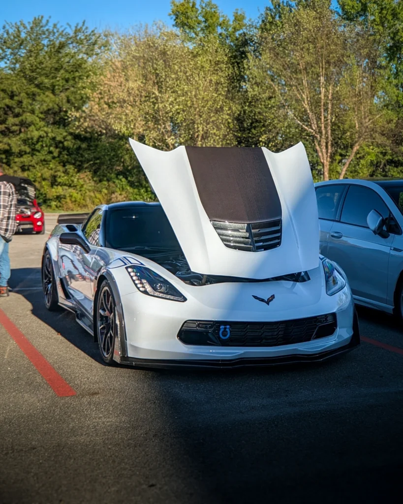 White Chevrolet Corvette with open hood showing black vent and grille, parked in lot with trees and other vehicles