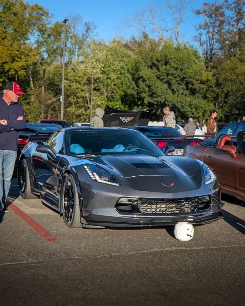 Black Chevrolet Corvette parked outside with a small white pumpkin in front and people standing nearby in a parking lot