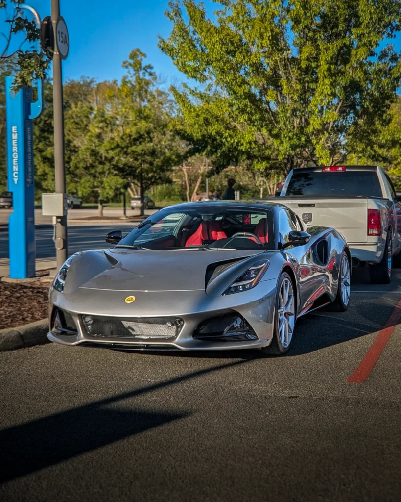 A silver Lotus Emira with red seats is parked beside a curb in a tree-lined parking lot.