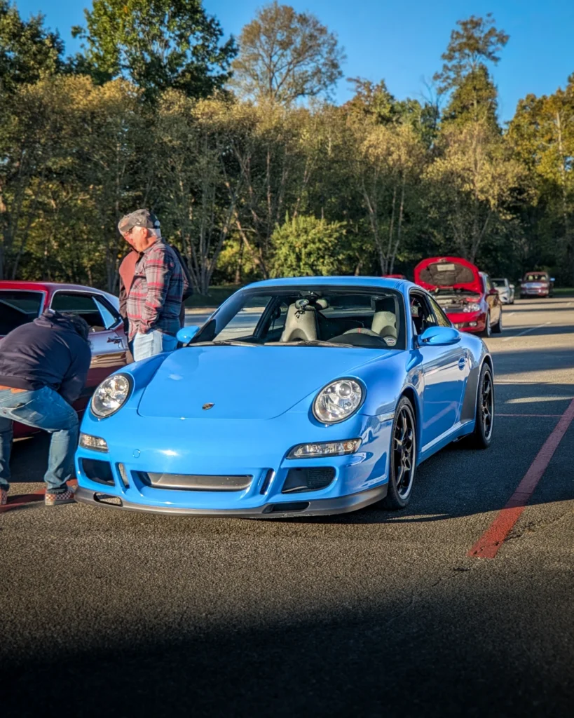 Blue Porsche 911 GT3 parked in a lot with people and other cars, trees in background under clear sky