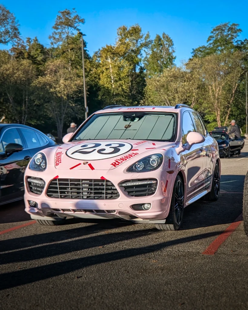 A pink Porsche Cayenne with number 23 on its hood is parked among other cars in a tree-lined lot.
